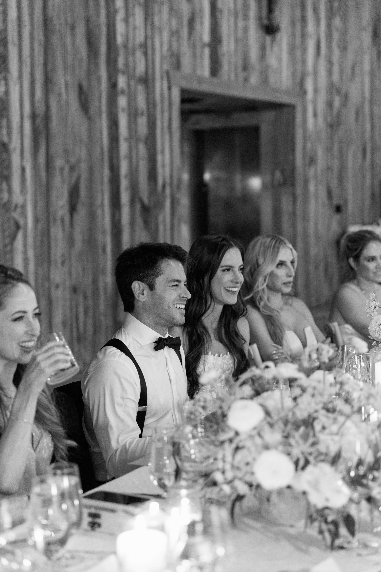 Wedding party seated at reception table with floral centerpiece and elegant decor in rustic barn setting.
