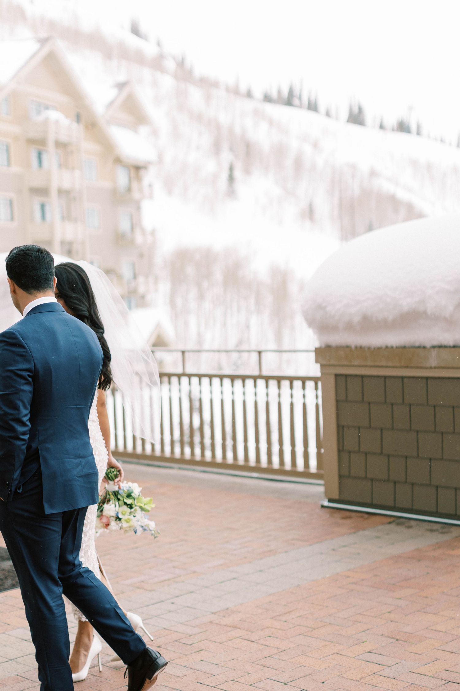 A wedding couple walks together on a snow-covered terrace with mountain lodges in the background.