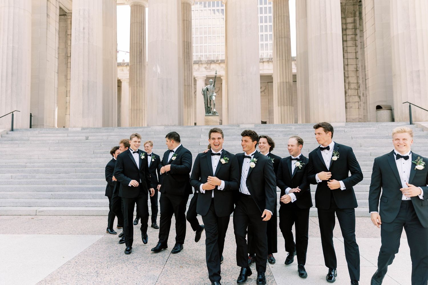 Groomsmen in black tuxedos gather on classical stone steps of building.