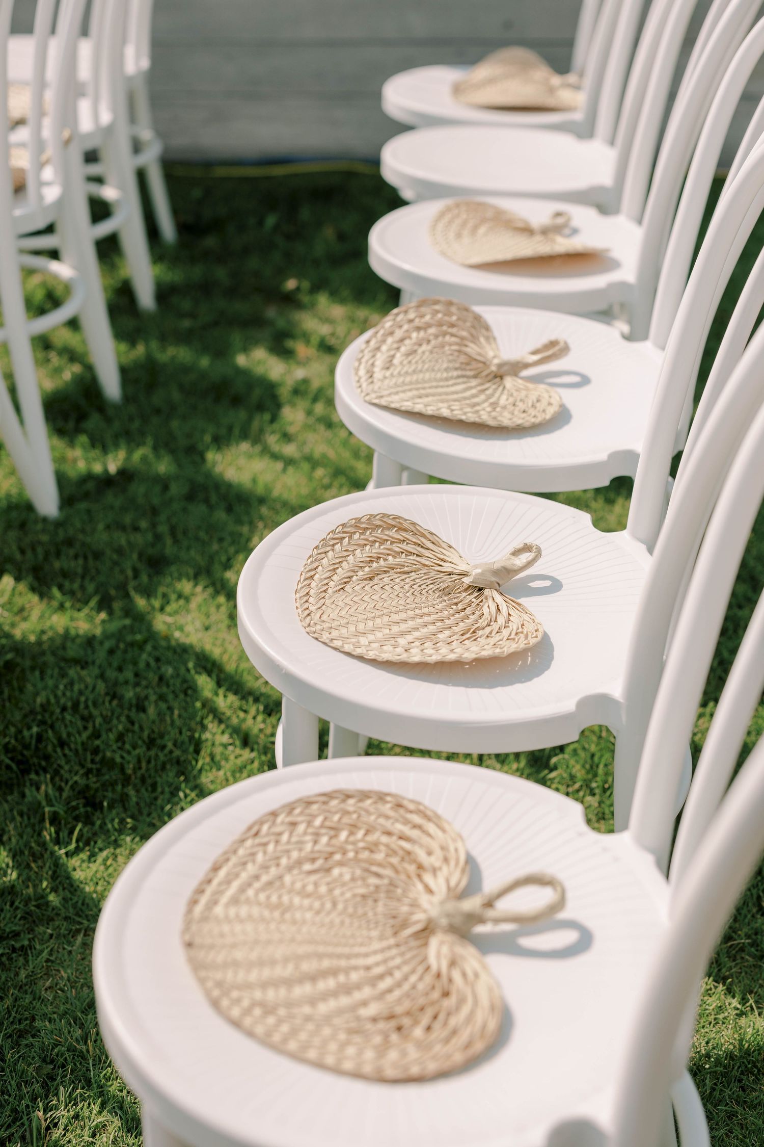 Row of white chairs with decorative woven palm leaf fans placed on each seat.