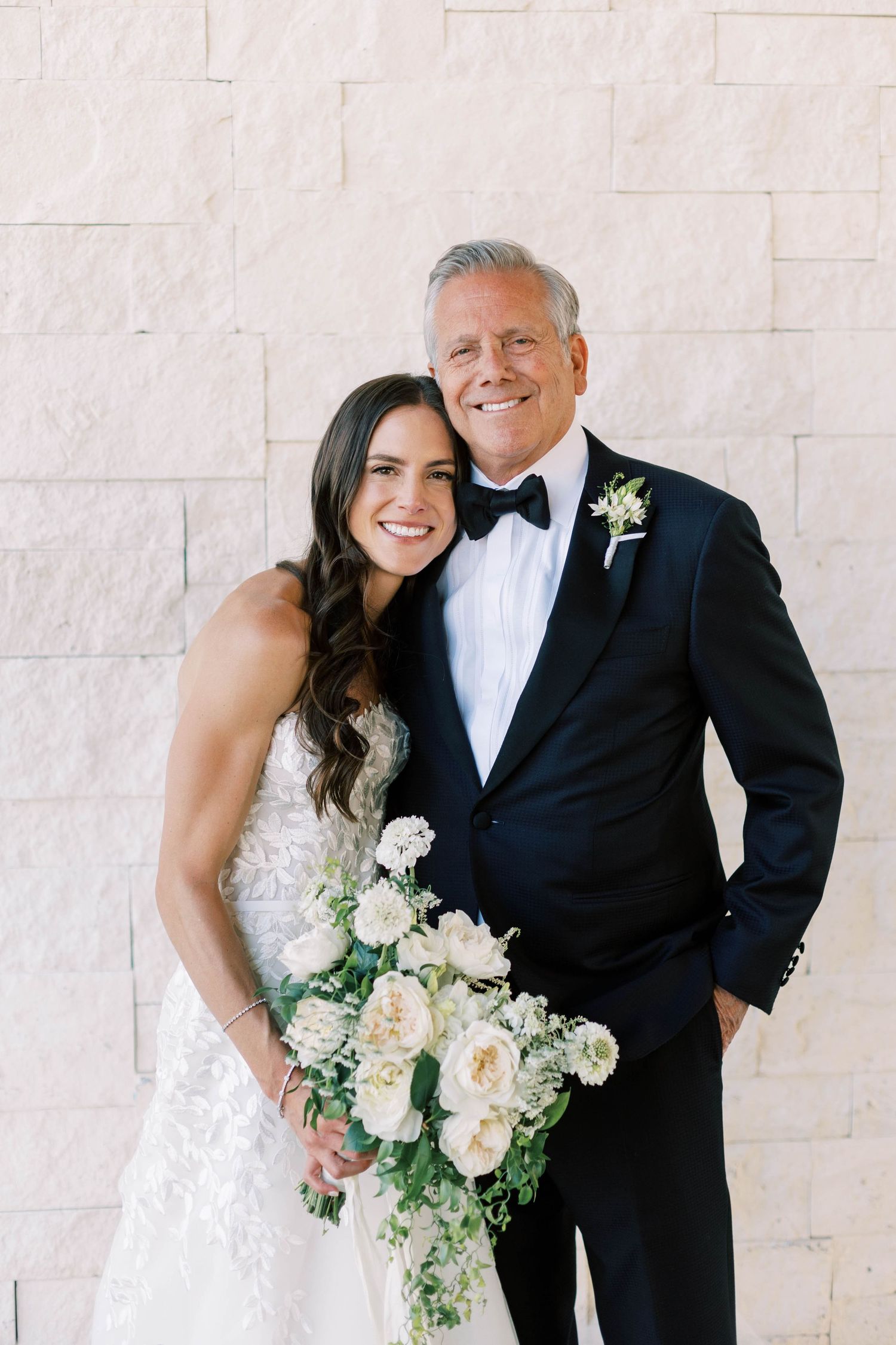 A wedding portrait featuring a bride in lace dress and groom in classic tuxedo against white stone wall.