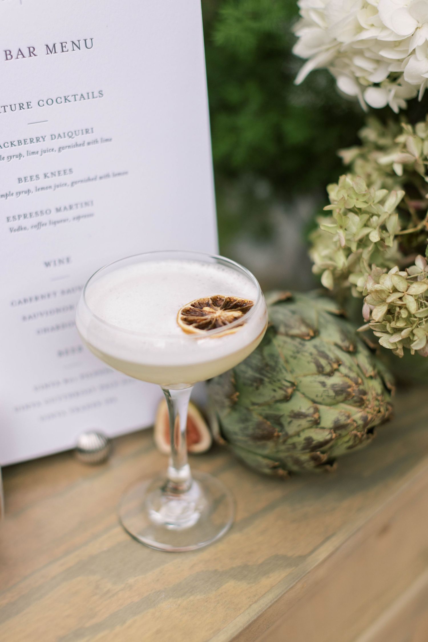 Elegant cocktail with frothy top and dried citrus garnish next to bar menu and floral decorations.
