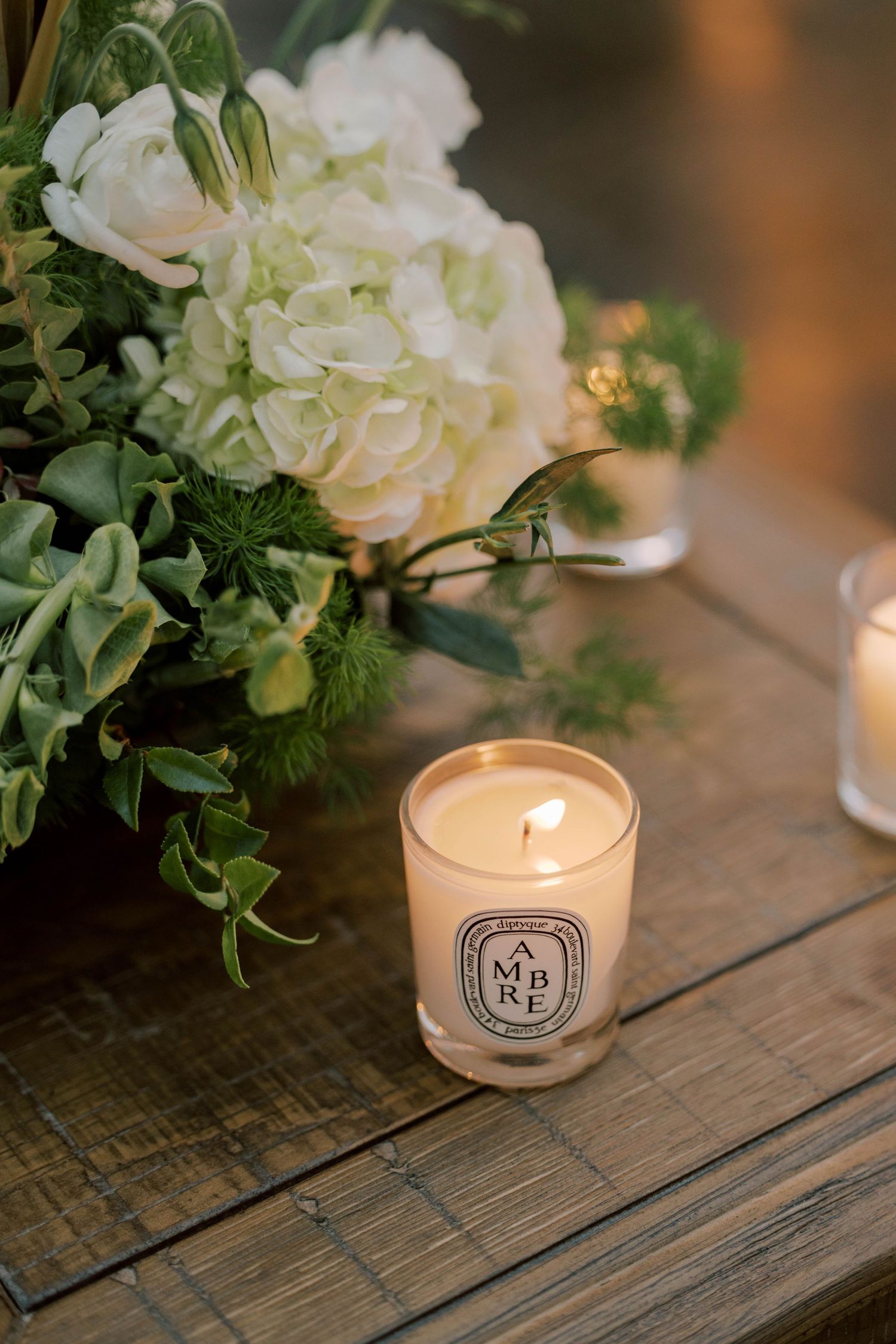 Elegant white floral arrangement with greenery next to lit Diptyque candle on wooden surface.