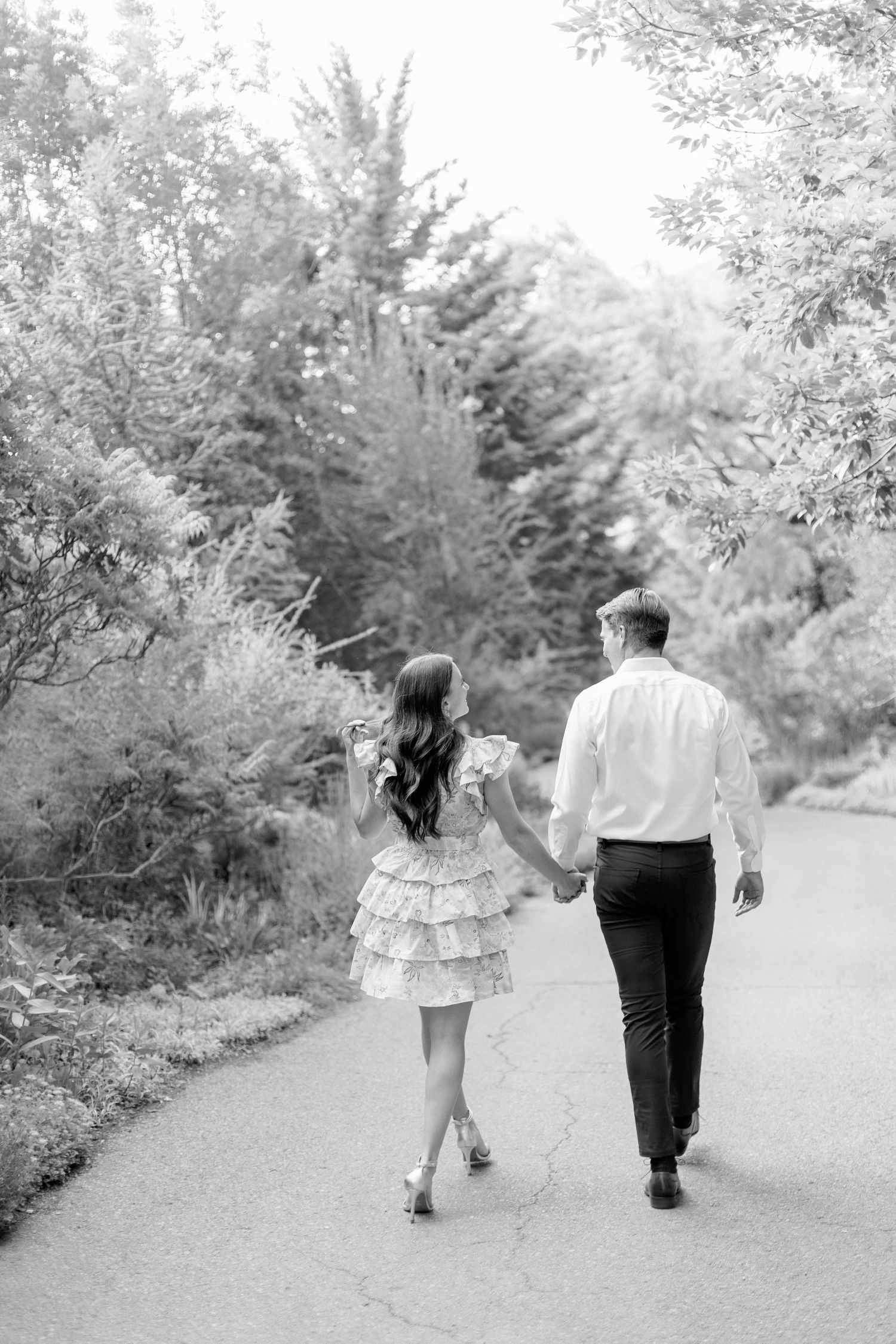 A black and white photo of a couple walking hand in hand along a tree-lined path.