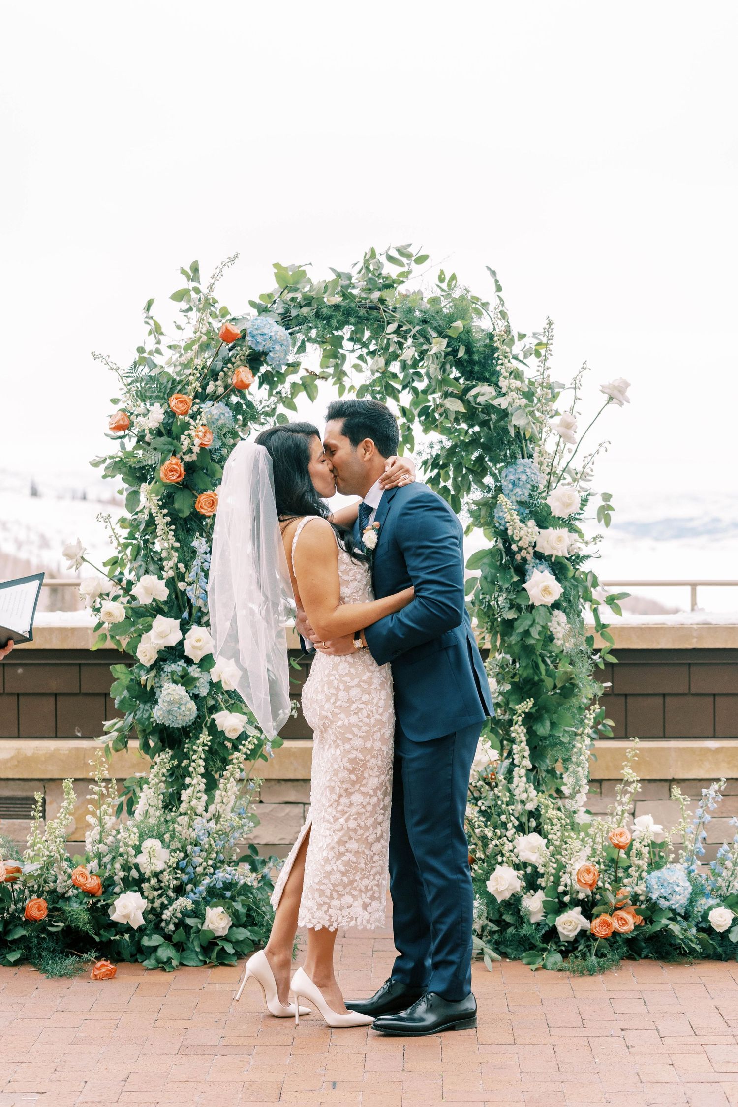 Couple sharing intimate moment under lush floral arch with white and peach blooms.