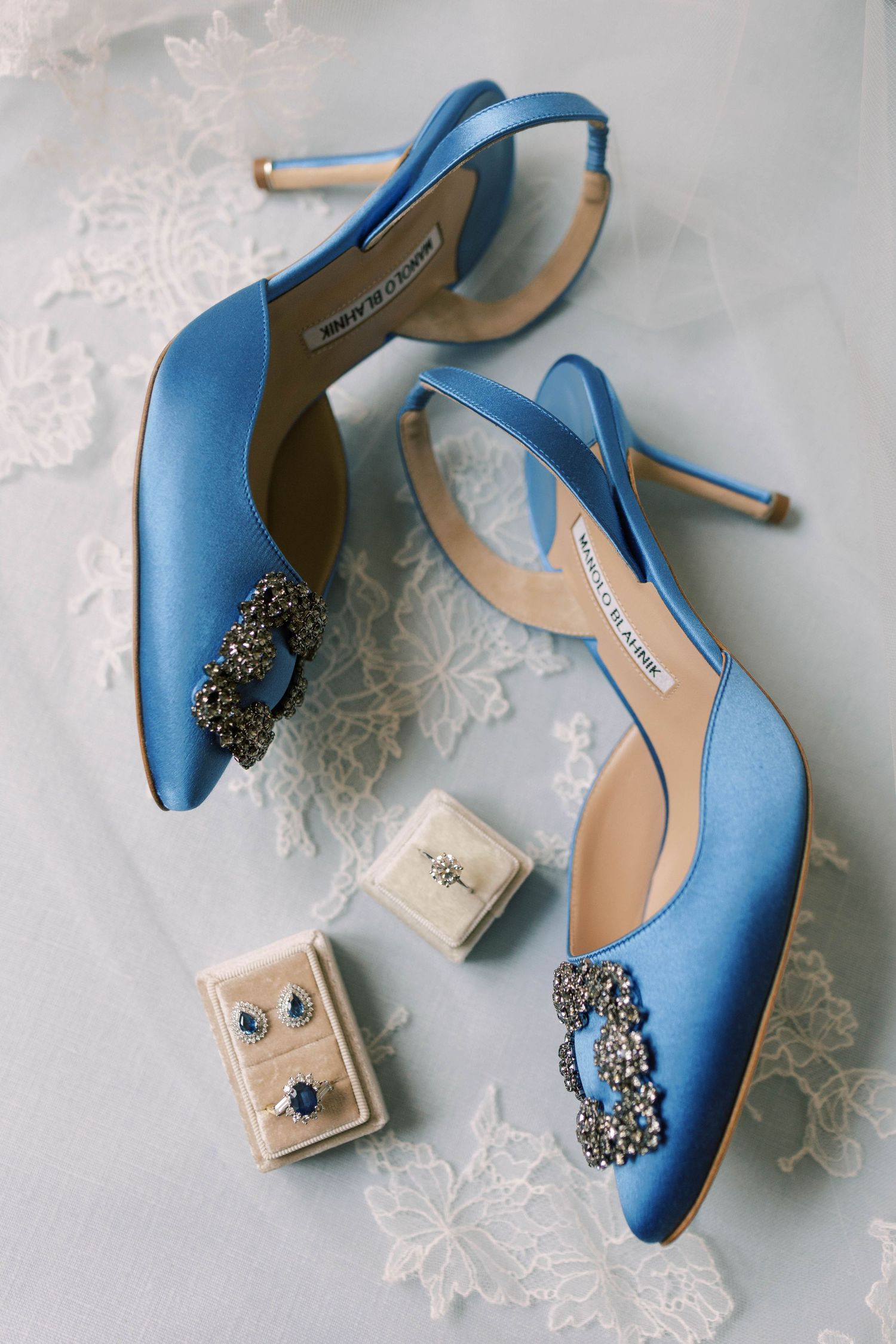 Elegant blue satin wedding shoes with crystal embellishments displayed with jewelry boxes on lace.