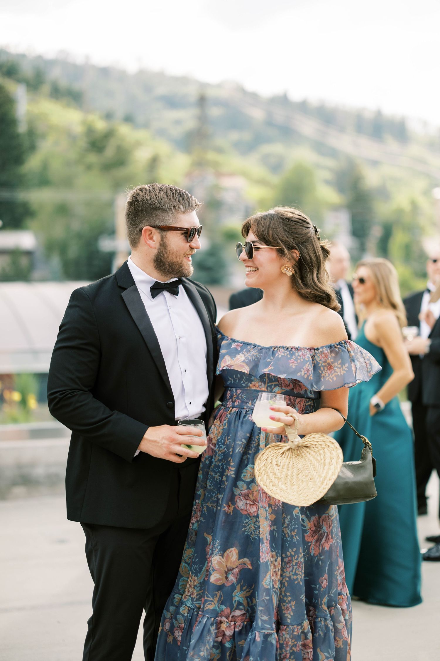 Couple in formal wear enjoying drinks at an outdoor event with mountain scenery.