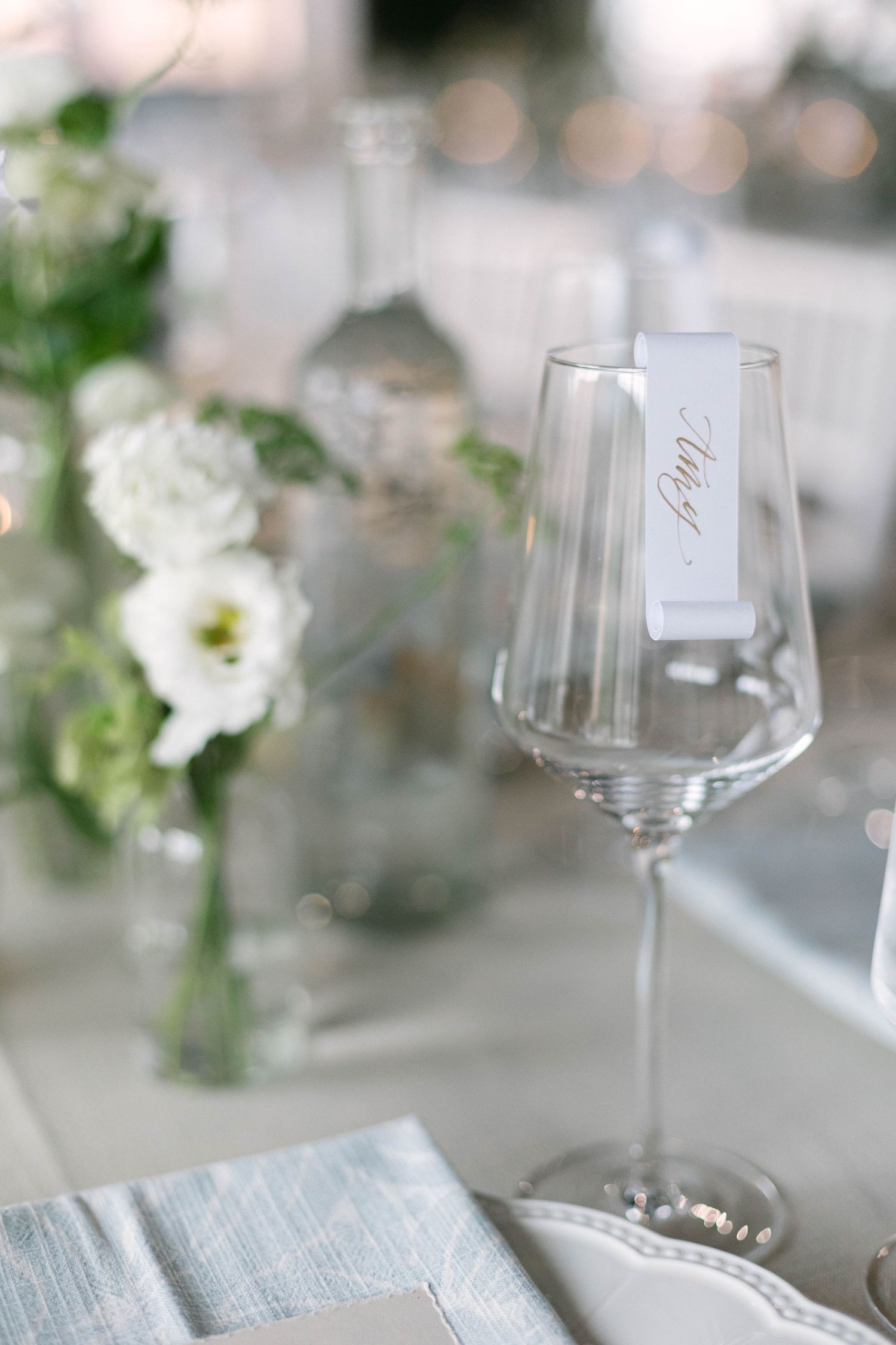 Elegant place setting with wine glass and name card alongside white floral arrangements on light tablecloth.