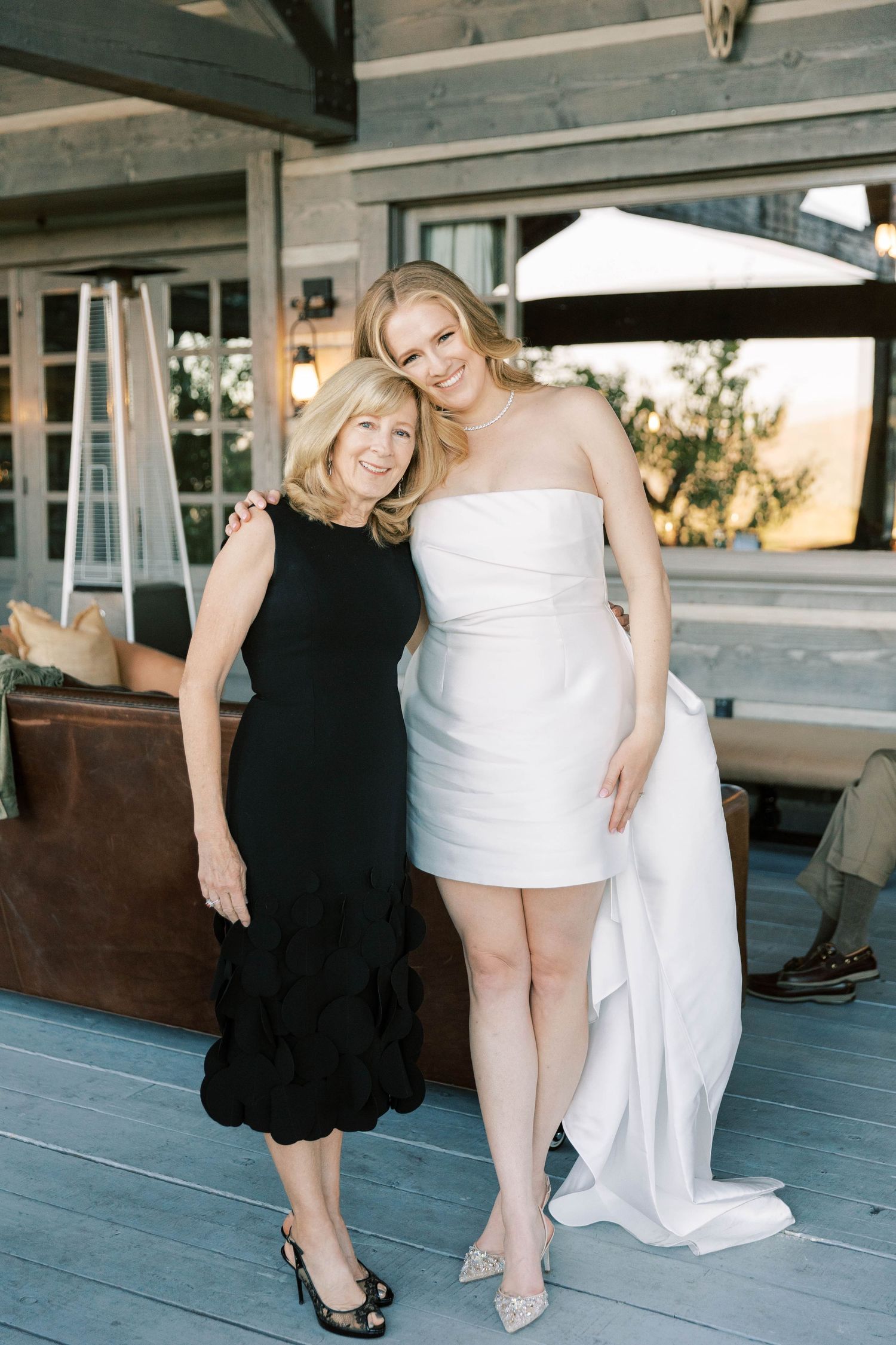 Two elegantly dressed women pose together on a rustic wooden deck during a formal event.