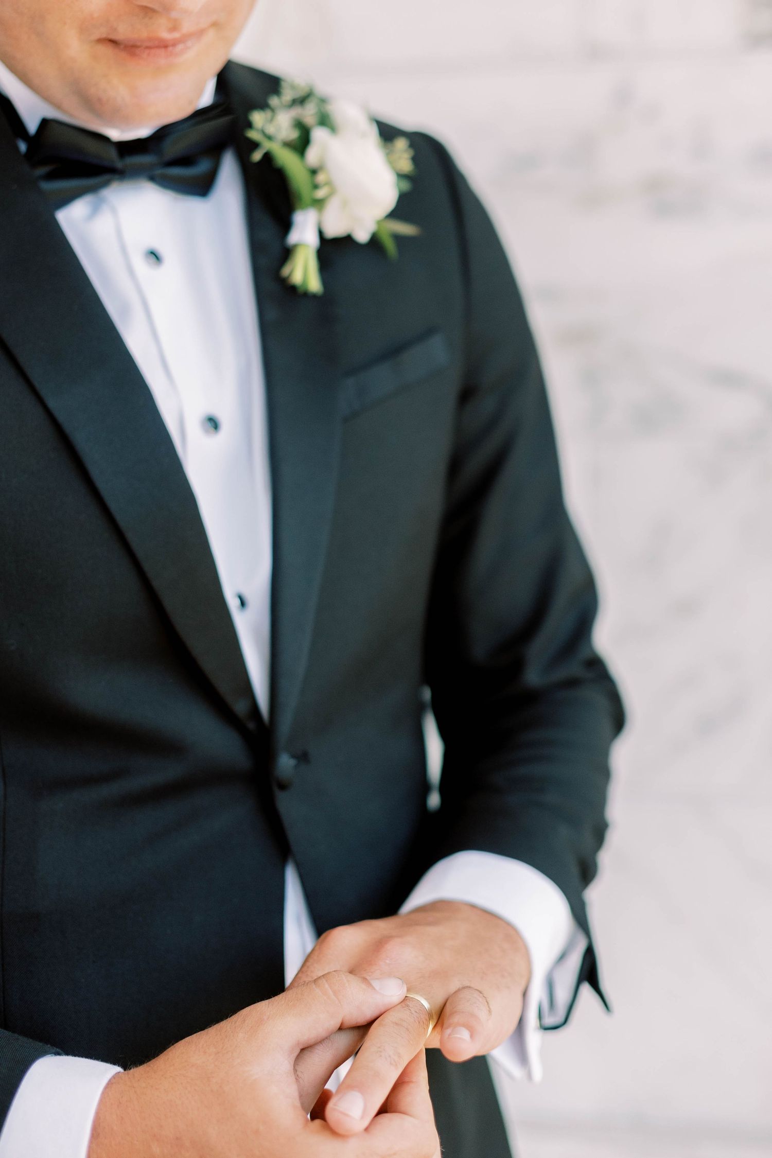 Close-up of groom in classic tuxedo with white boutonniere and wedding ring.