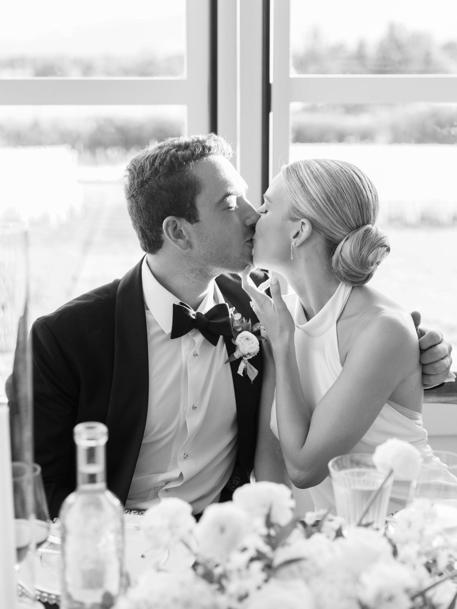 A romantic kiss between newlyweds at their wedding reception table with champagne and floral decorations.