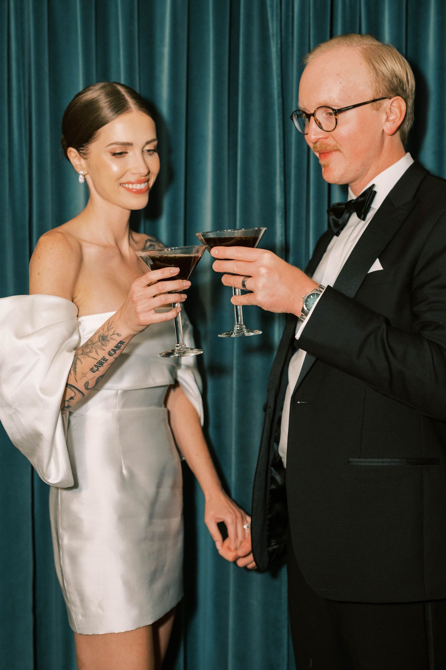 Couple in formal attire sharing cocktails against teal curtain backdrop.