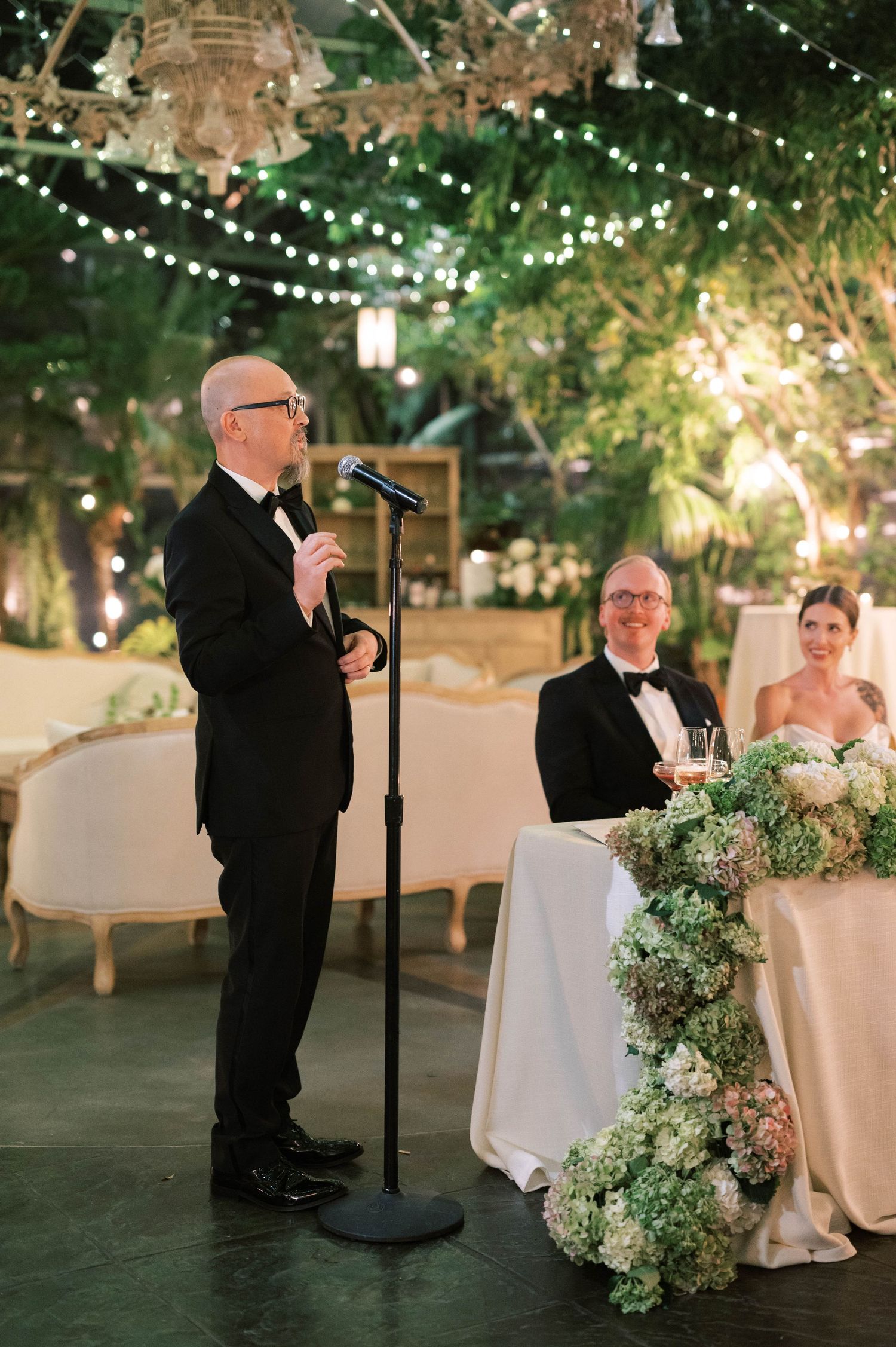 Formal speech being delivered during wedding reception under string lights and chandeliers.