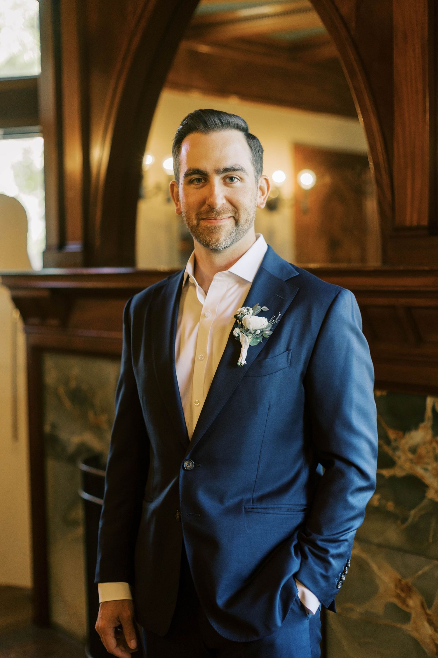 Wedding attire featuring a navy suit with boutonniere and pink dress shirt in an elegant indoor setting.
