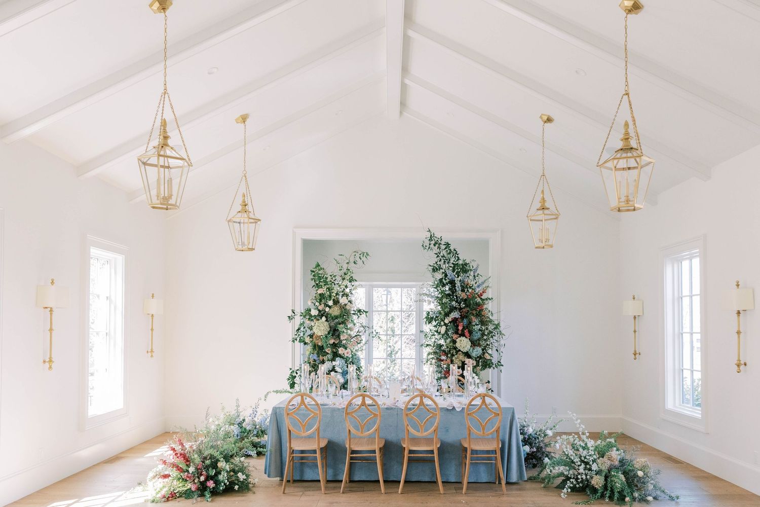 Light-filled wedding reception room with vaulted ceiling, gold lanterns, and blue table settings.