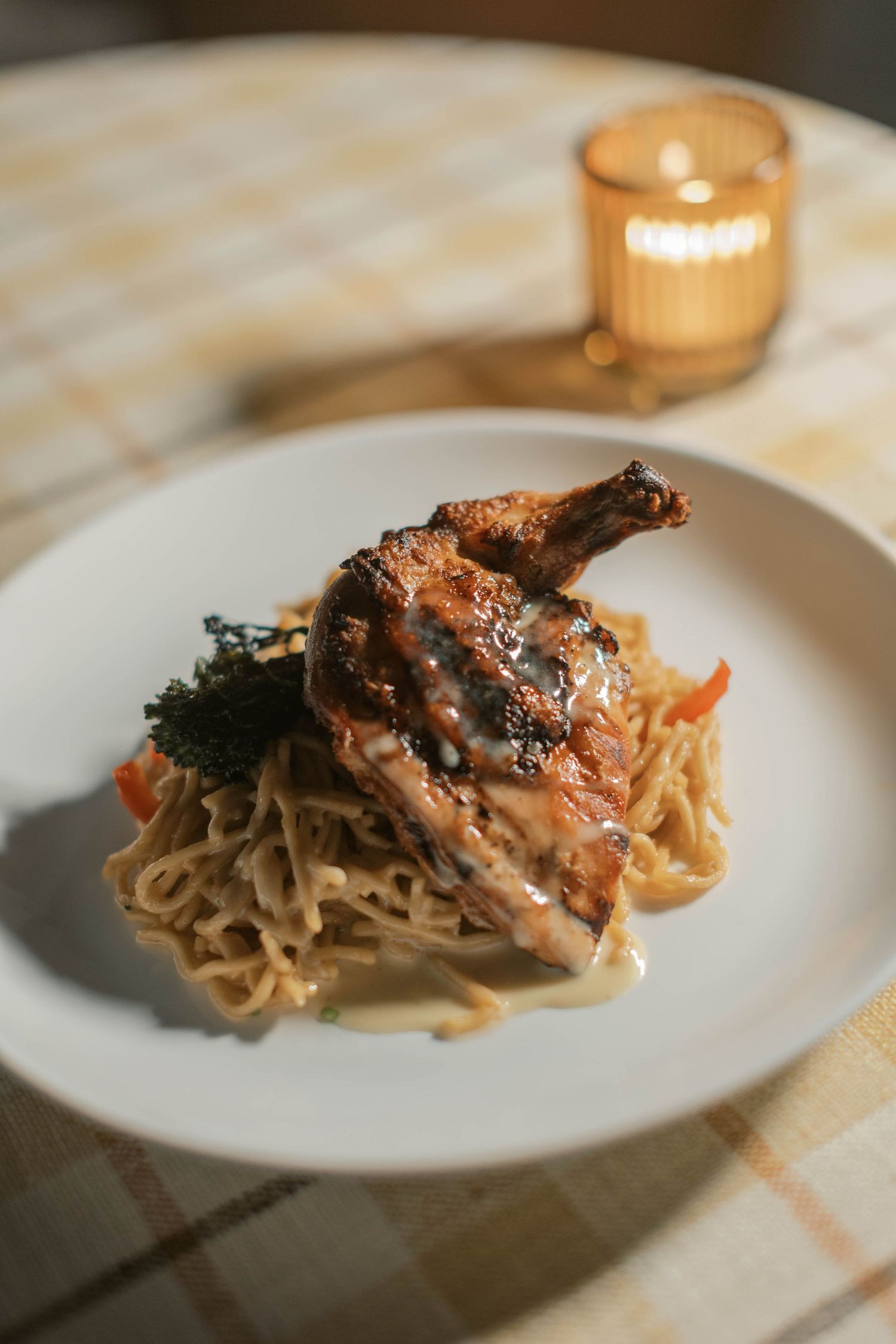 A plate of roasted chicken and pasta sits on a checkered tablecloth with a glowing candle in the background.