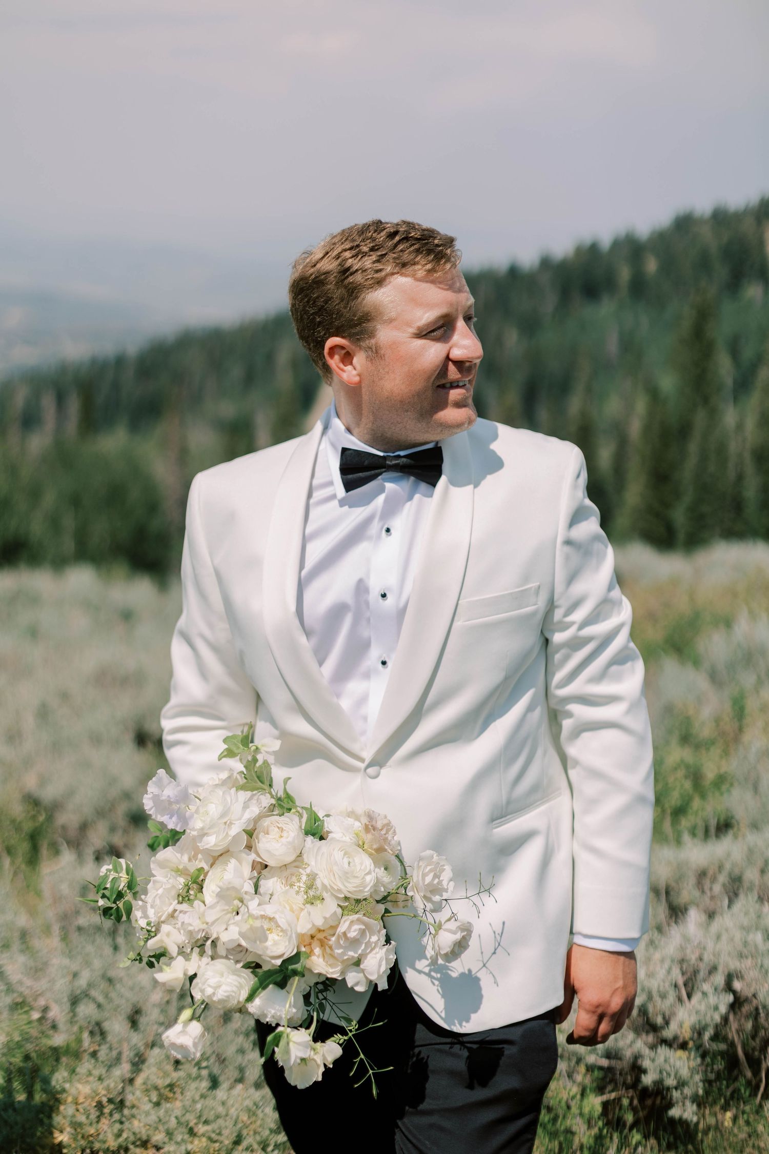 Groom in white tuxedo jacket holds a bouquet of white flowers against mountain landscape.