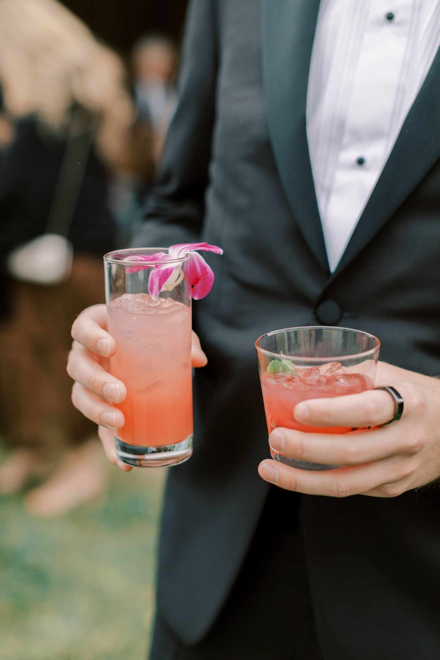 Person in tuxedo holding pink signature cocktails with ribbons at wedding reception.