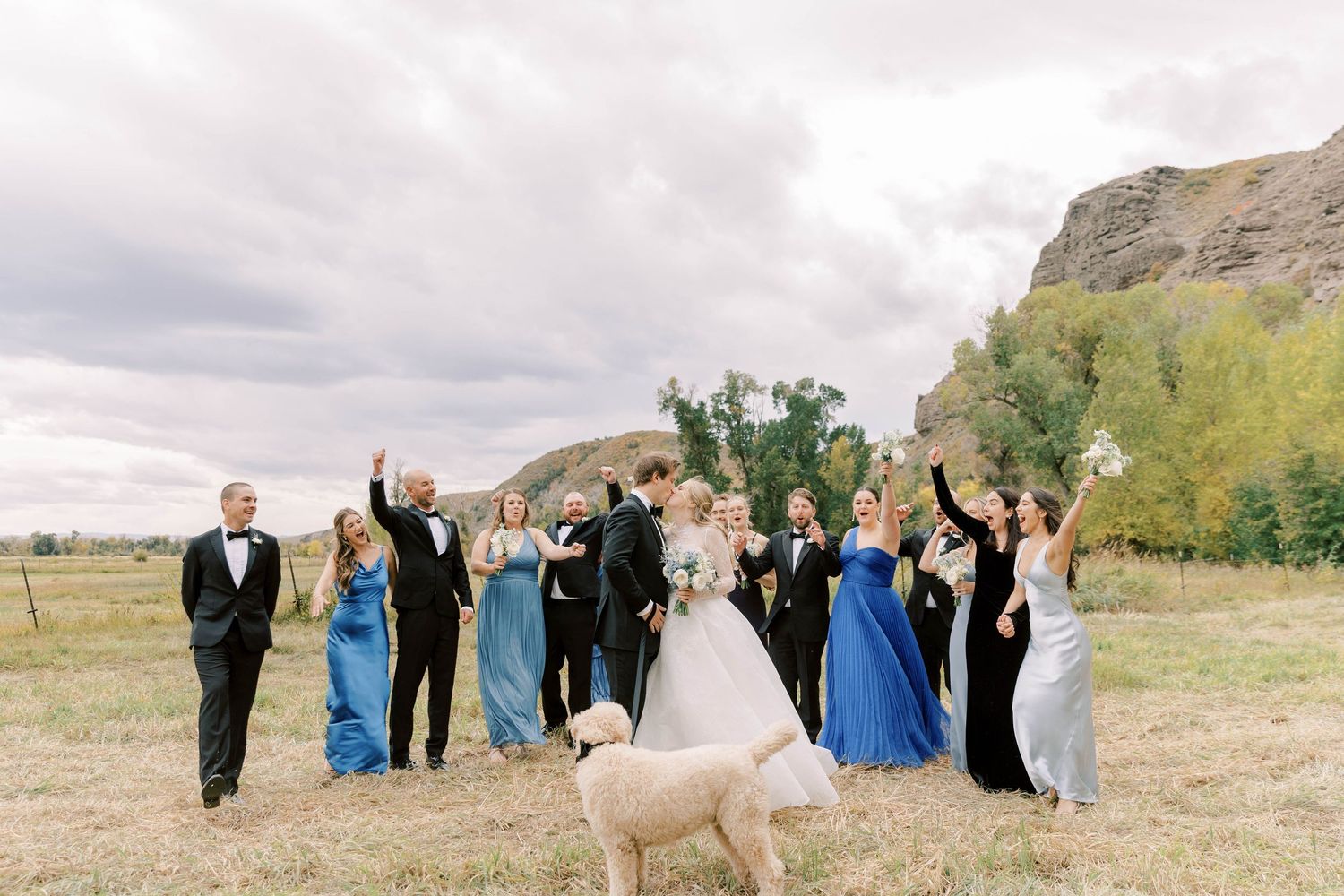 Wedding party in formal attire poses outdoors with fluffy white dog in foreground.