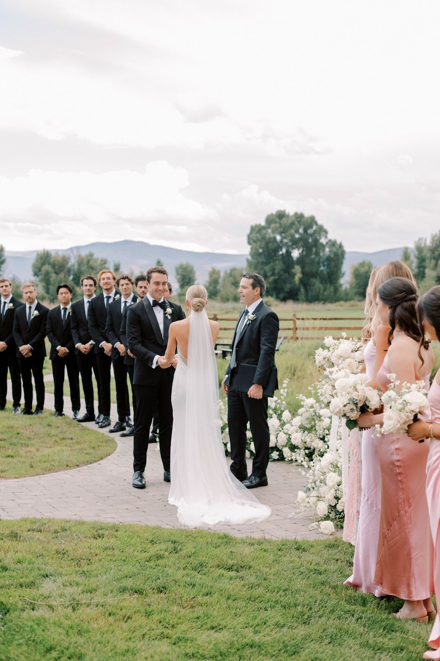 Wedding party lined up along flower-decorated path with mountain backdrop during outdoor ceremony.