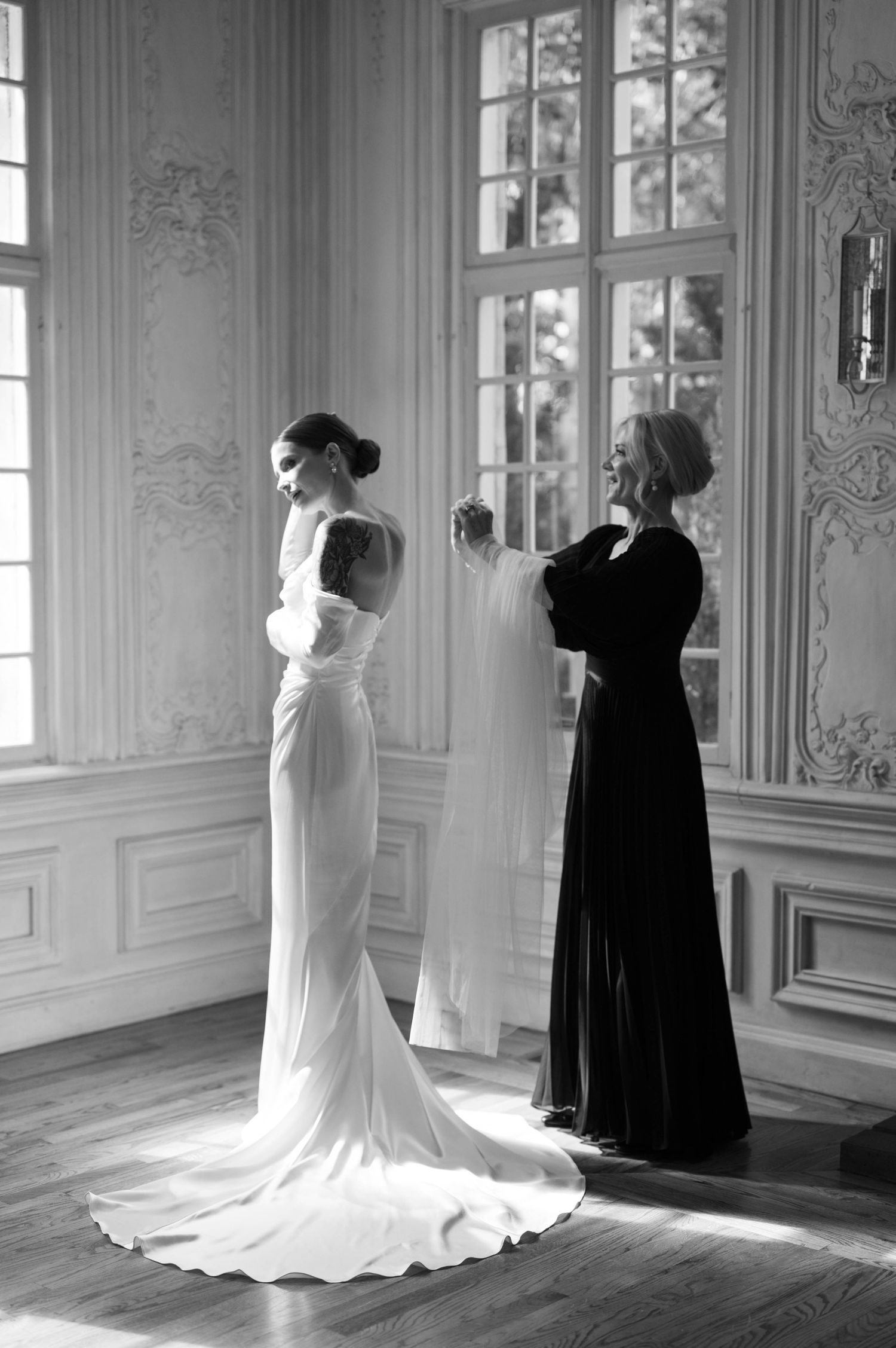 Two elegant gowns side by side in an ornate room with high windows and decorative molding details.