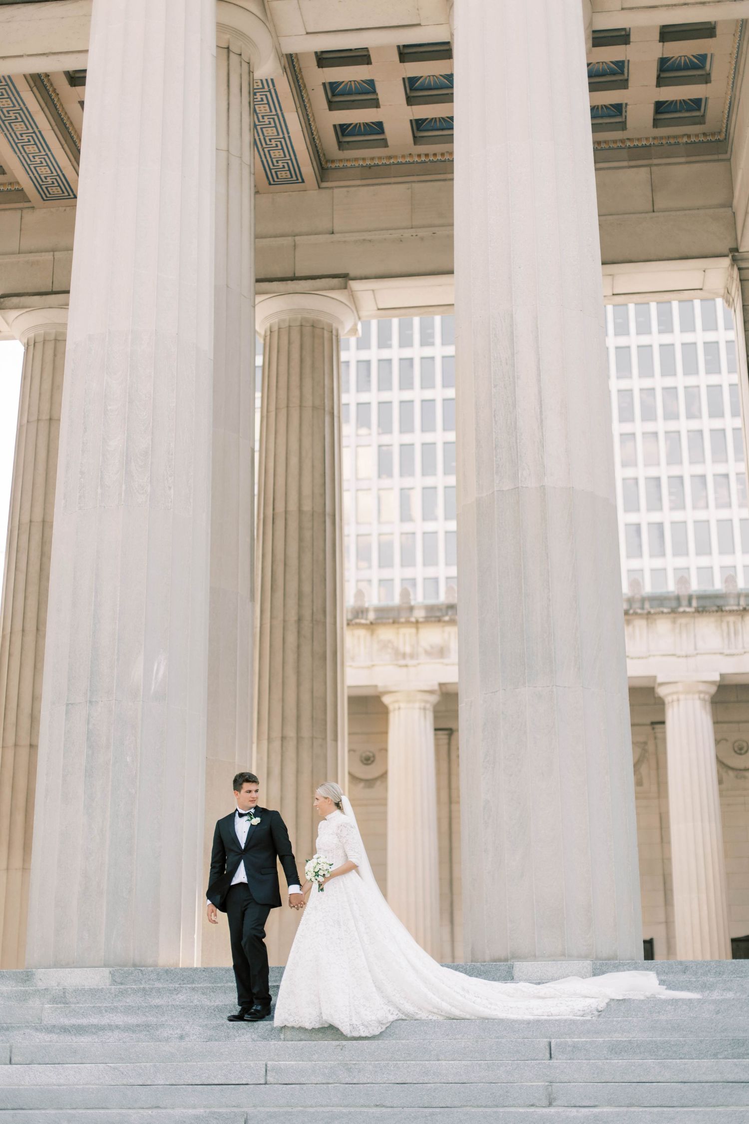 Wedding couple standing between grand classical columns of historic building with trailing bridal gown.
