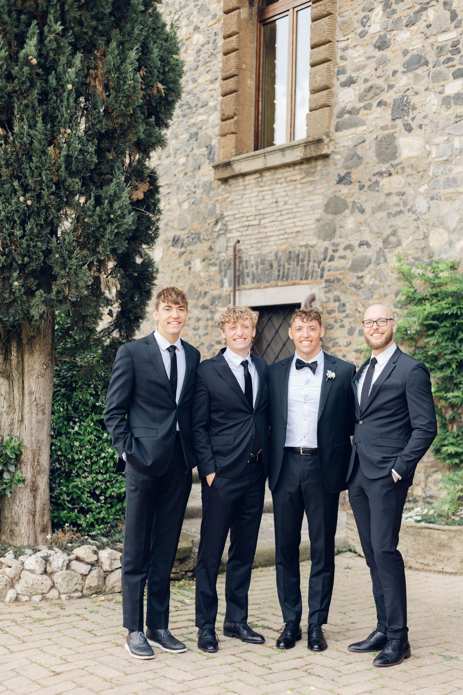 Four men in dark suits pose together against stone villa exterior.