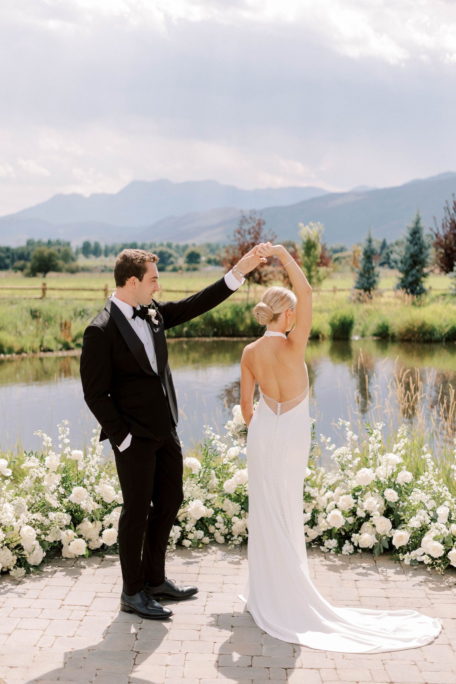 Couple shares romantic dance moment by pond with mountain backdrop and white flowers.