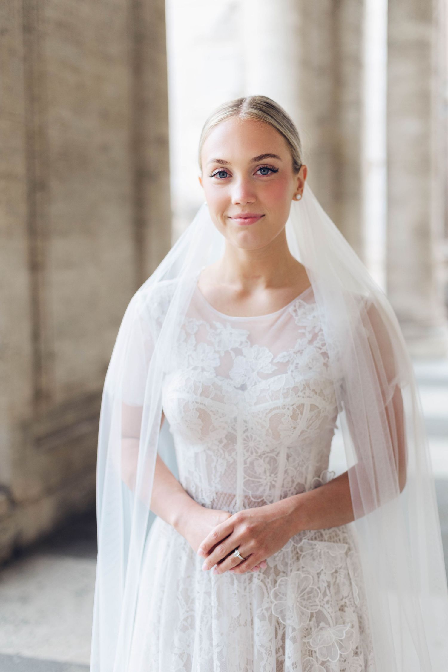 A bride wearing a white lace gown and veil poses in a classical stone setting of rome italy.
