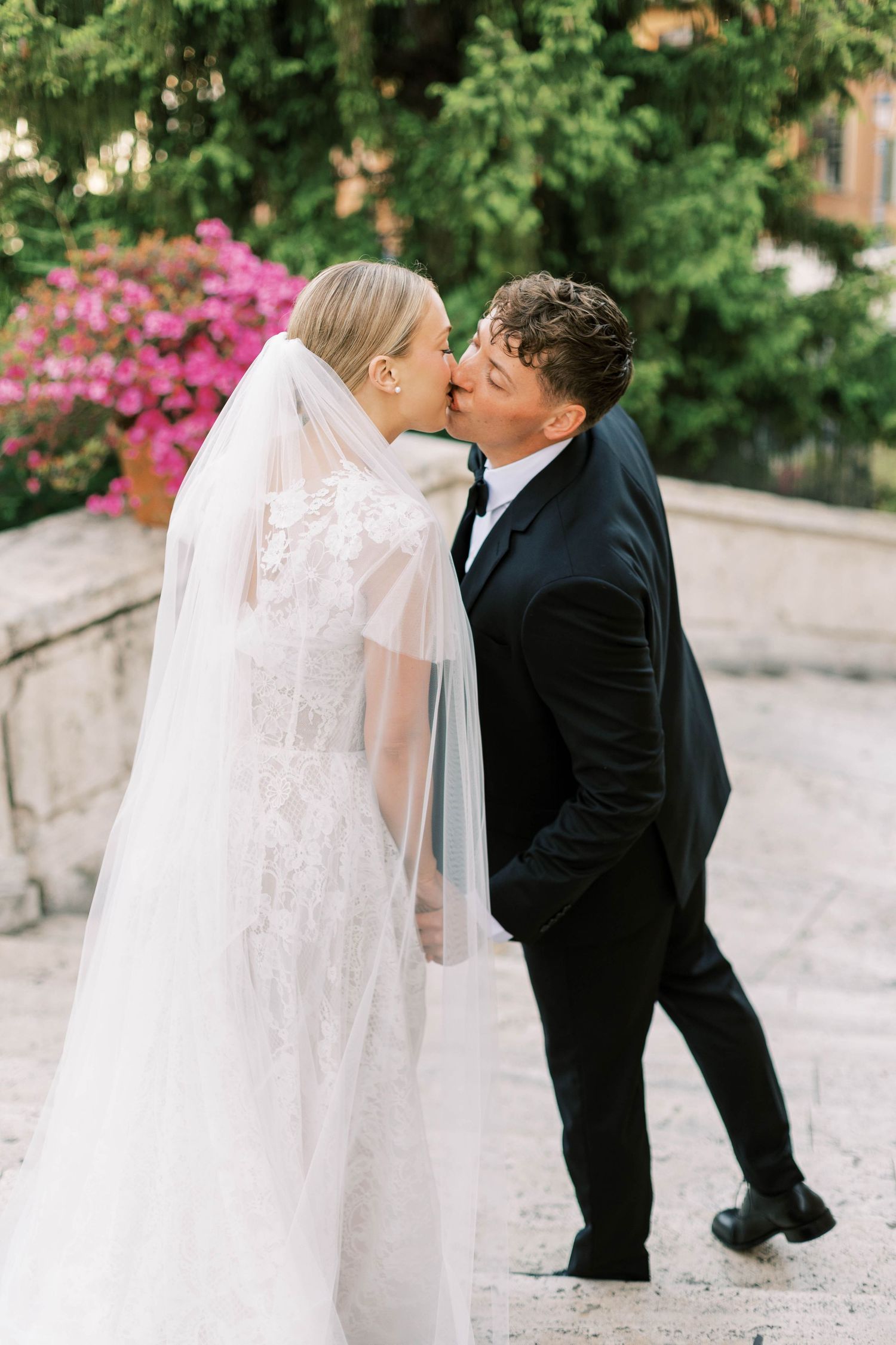 A tender wedding kiss moment captured in a garden setting with pink flowers and greenery.