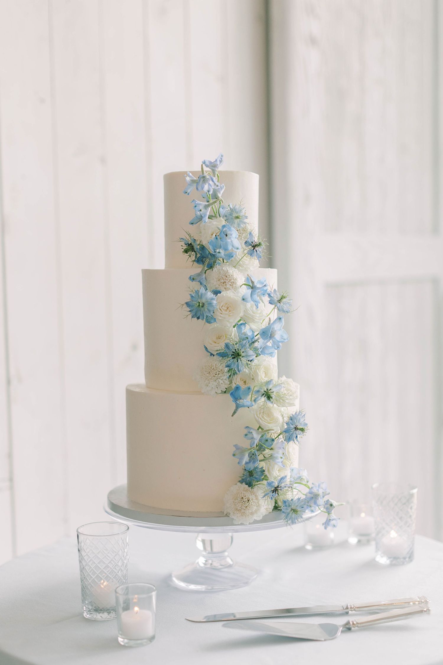 Three-tiered white wedding cake decorated with cascading blue and white flowers.