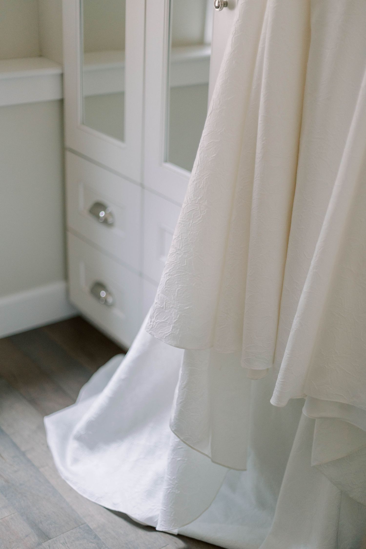Close-up of a white wedding dress hem against white cabinetry and wooden flooring.