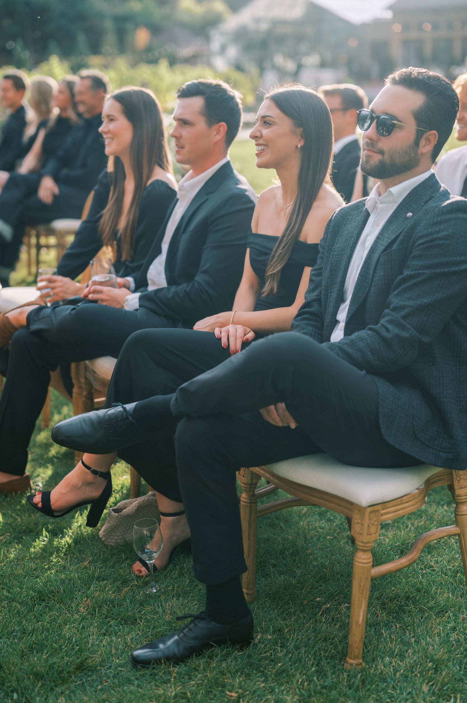 Wedding guests in formal attire seated on wooden chairs during outdoor ceremony.