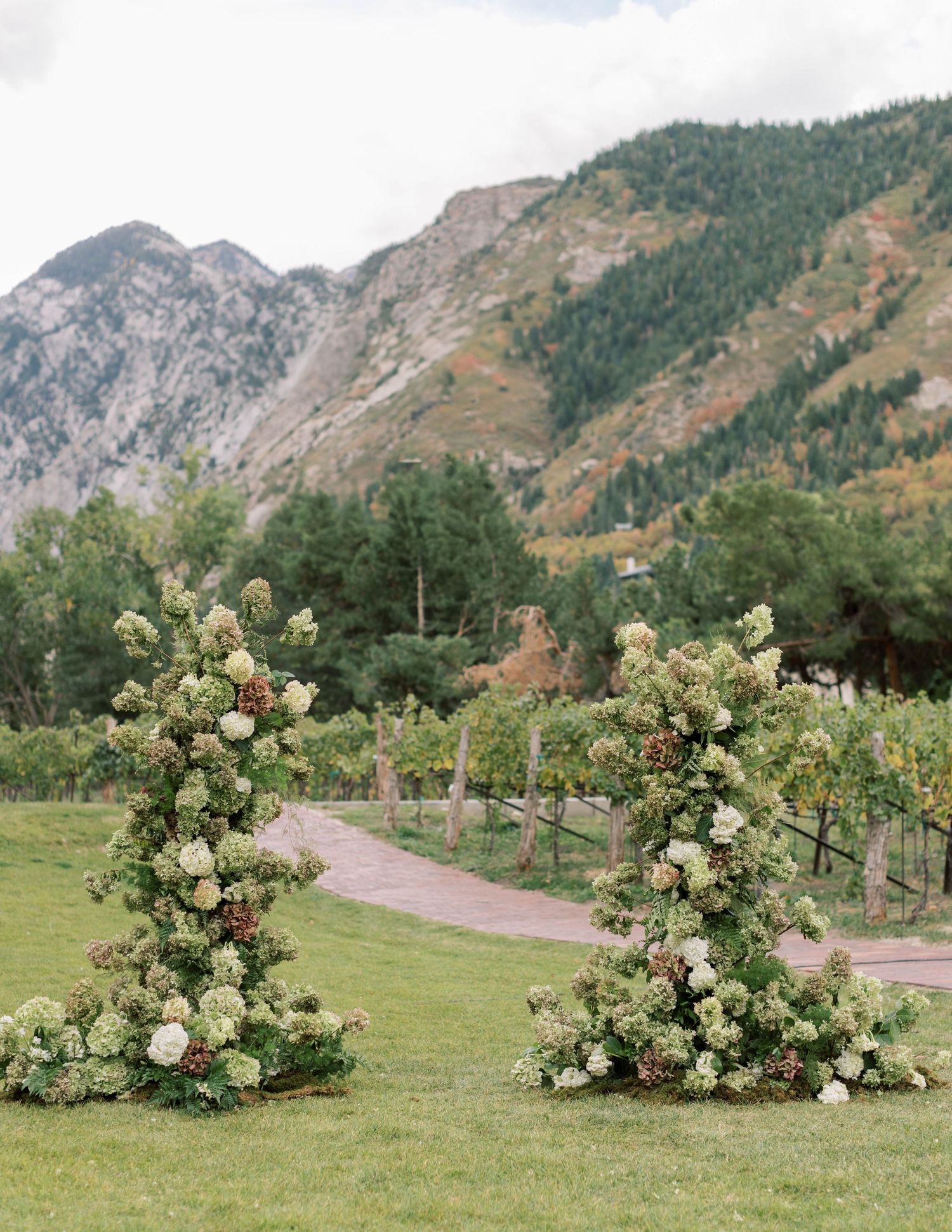 White floral archway frames a path with dramatic mountain backdrop in an outdoor wedding ceremony setting.