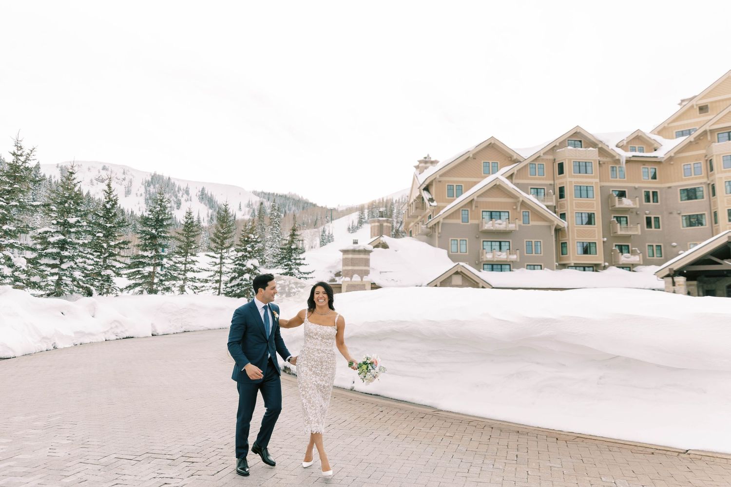 A winter wedding couple walking along a snowy path at montage deer valley with rustic mountain lodges in the background.
