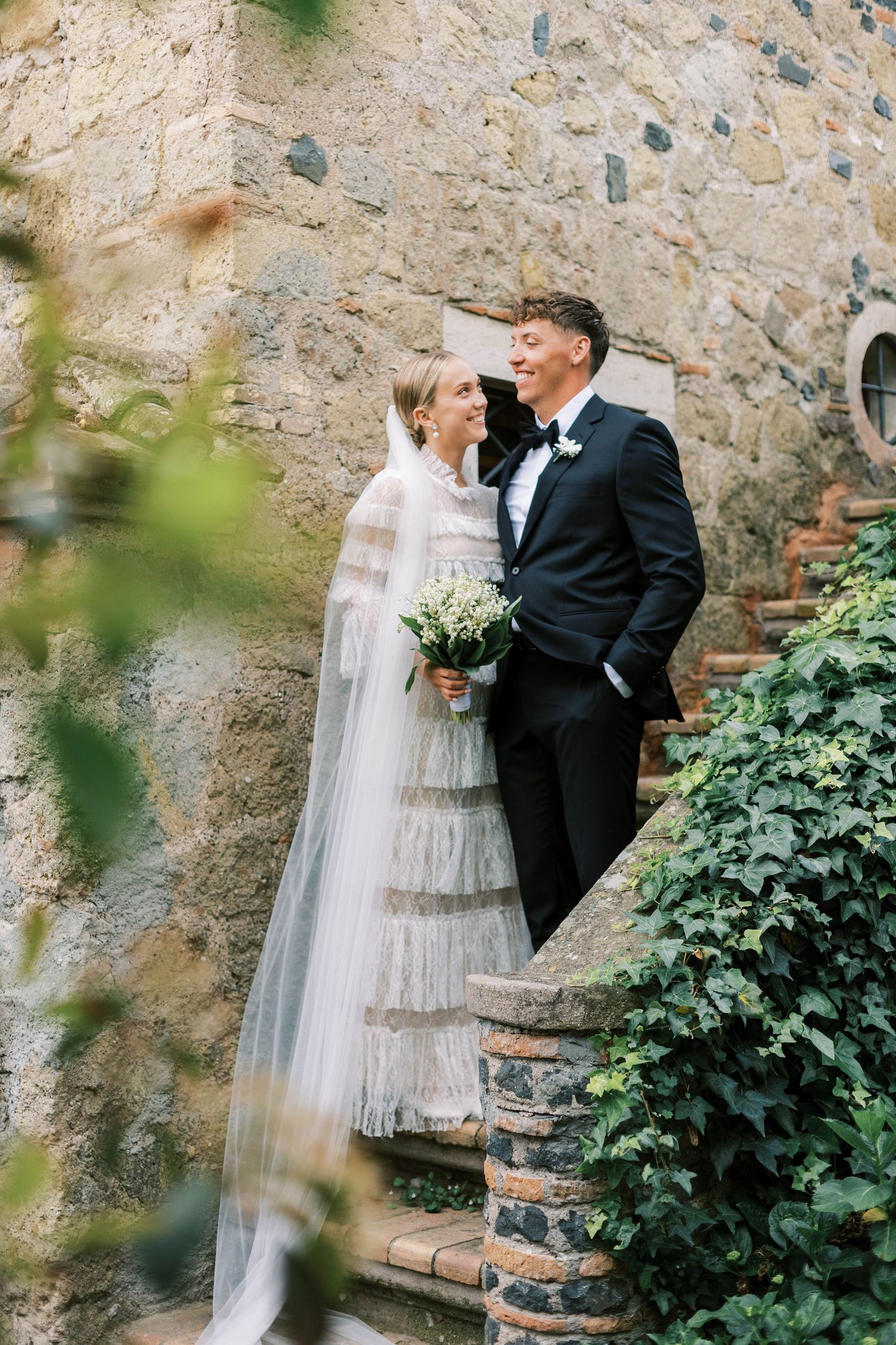 Wedding couple embracing on stone steps against rustic wall with greenery at castello della castellucia in rome italy.