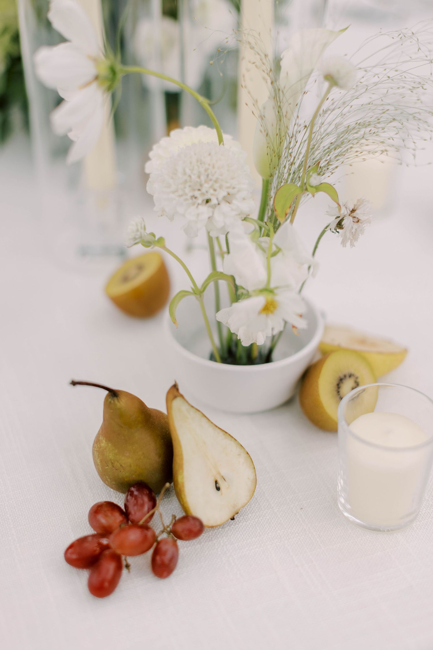 Still life arrangement of white flowers, pears, and red grapes on white tablecloth.