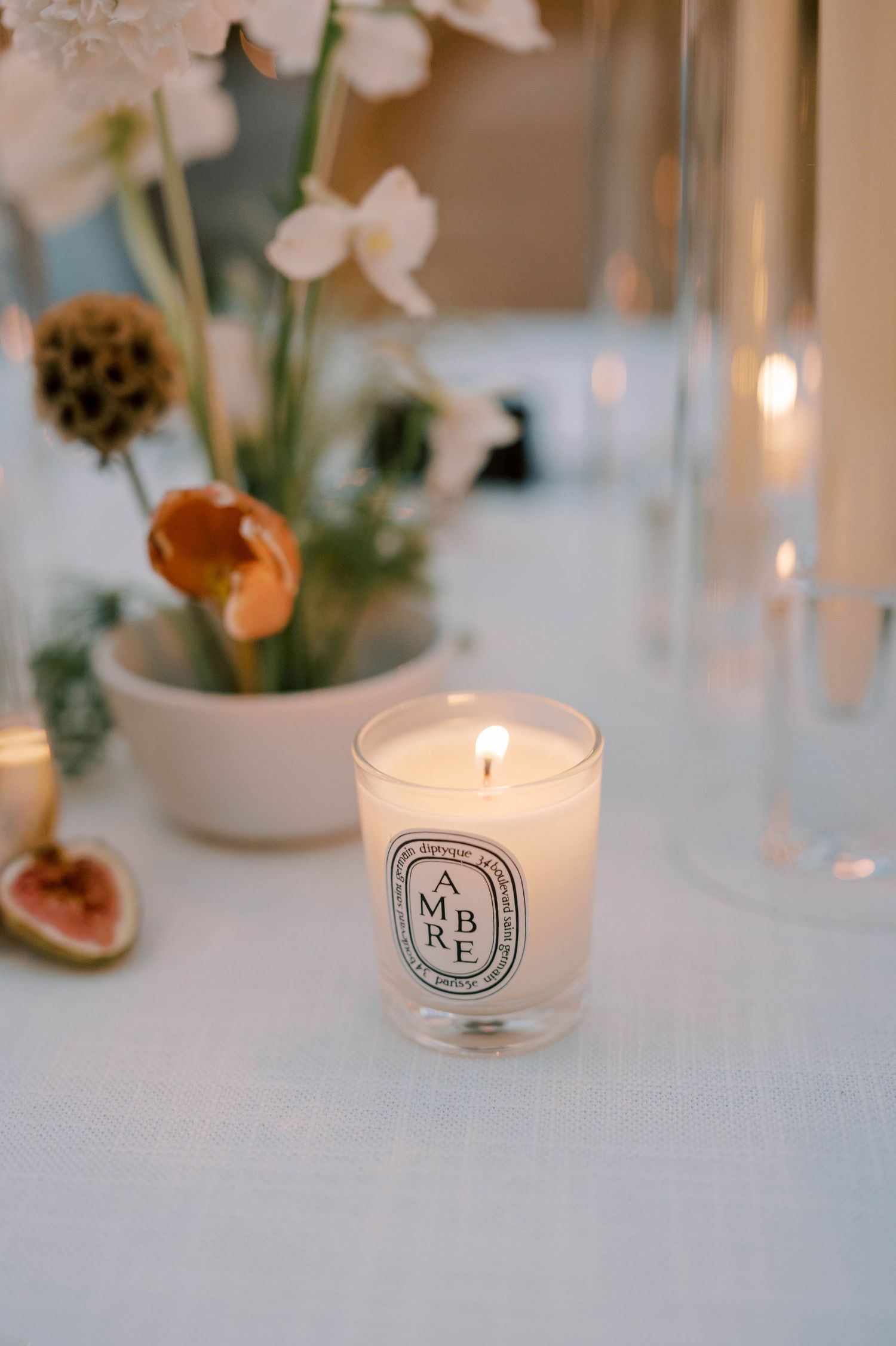 Diptyque candle and delicate floral arrangement on white tablecloth with soft lighting.