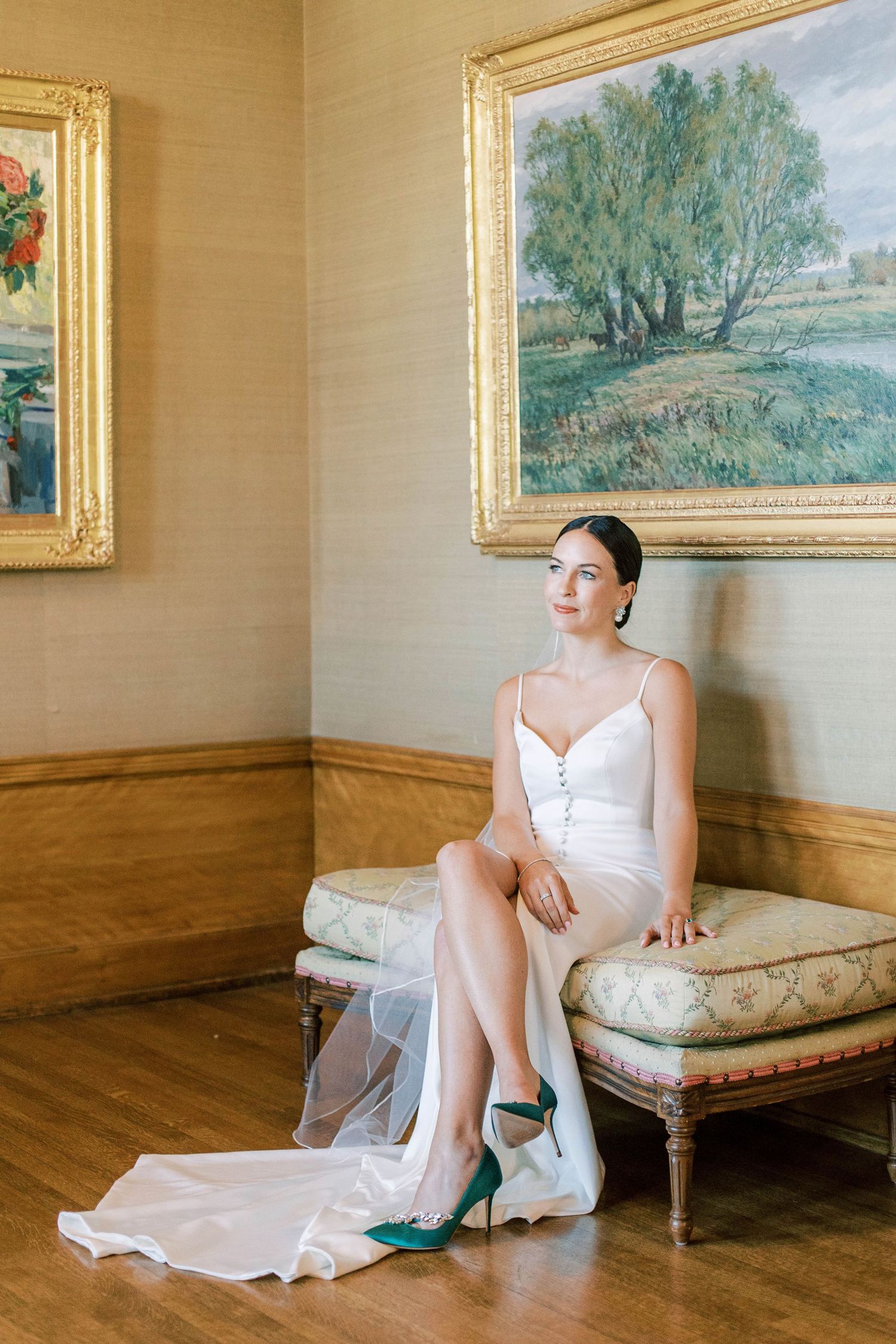 Bride in white dress sitting elegantly on antique bench between gilded frame paintings.