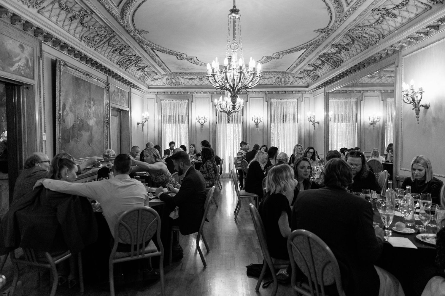Elegant black and white view of a formal dining room with ornate ceiling details and chandeliers.