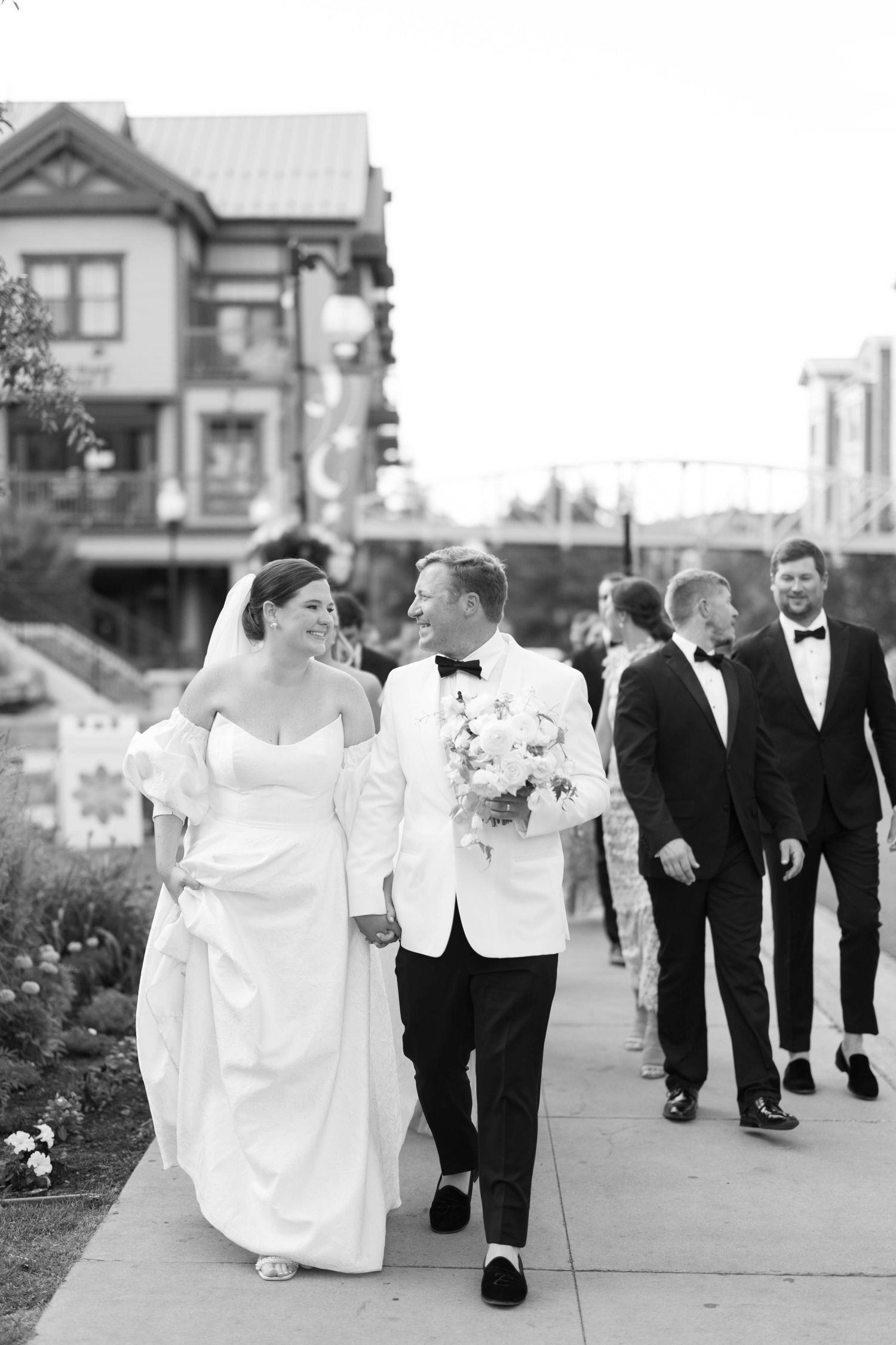Wedding party walking down sidewalk in formal attire with bride in white dress and groom in white jacket.