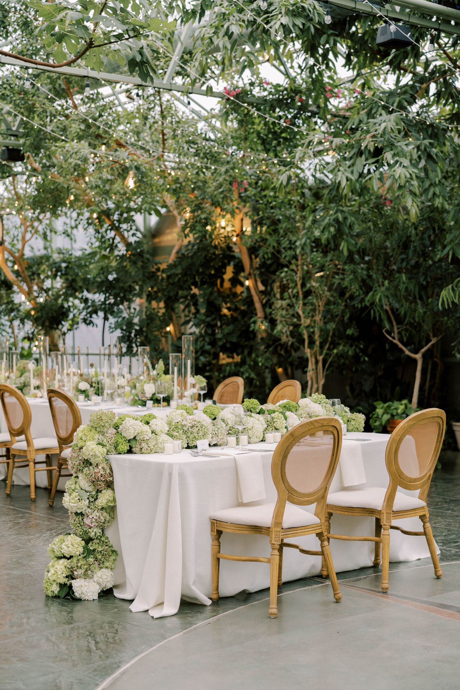 Greenhouse wedding reception with white tablecloths, French chairs, and lush hydrangea arrangements.