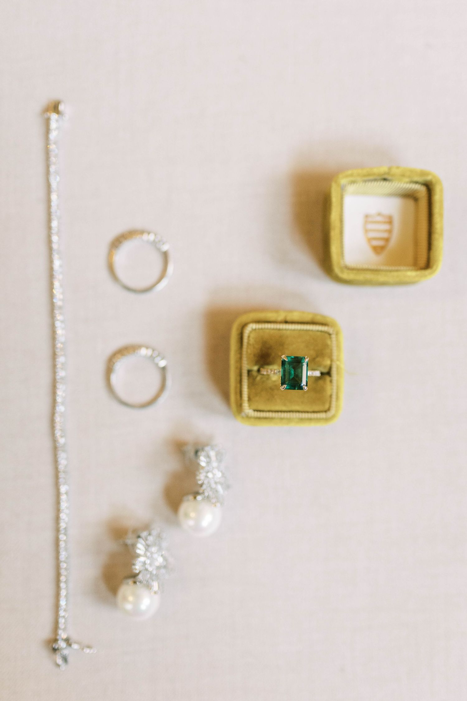 Wedding jewelry including rings, earrings and emerald engagement ring arranged on beige velvet boxes.