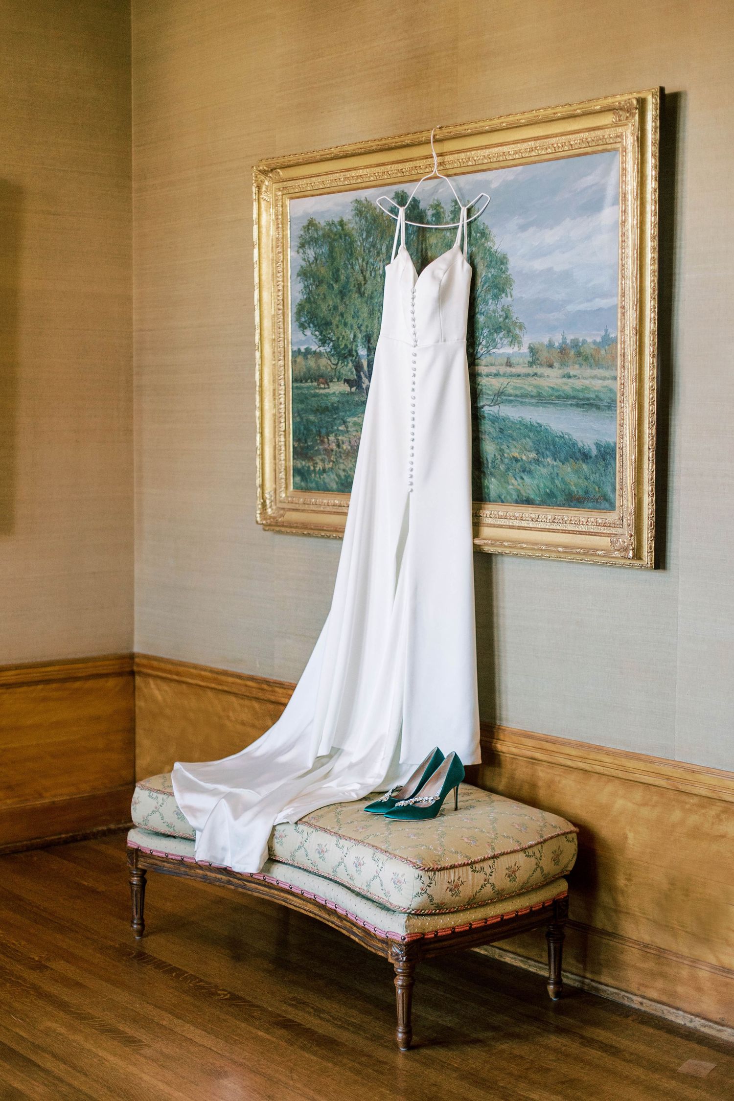 Elegant white wedding dress and teal shoes displayed on antique bench with framed landscape artwork.