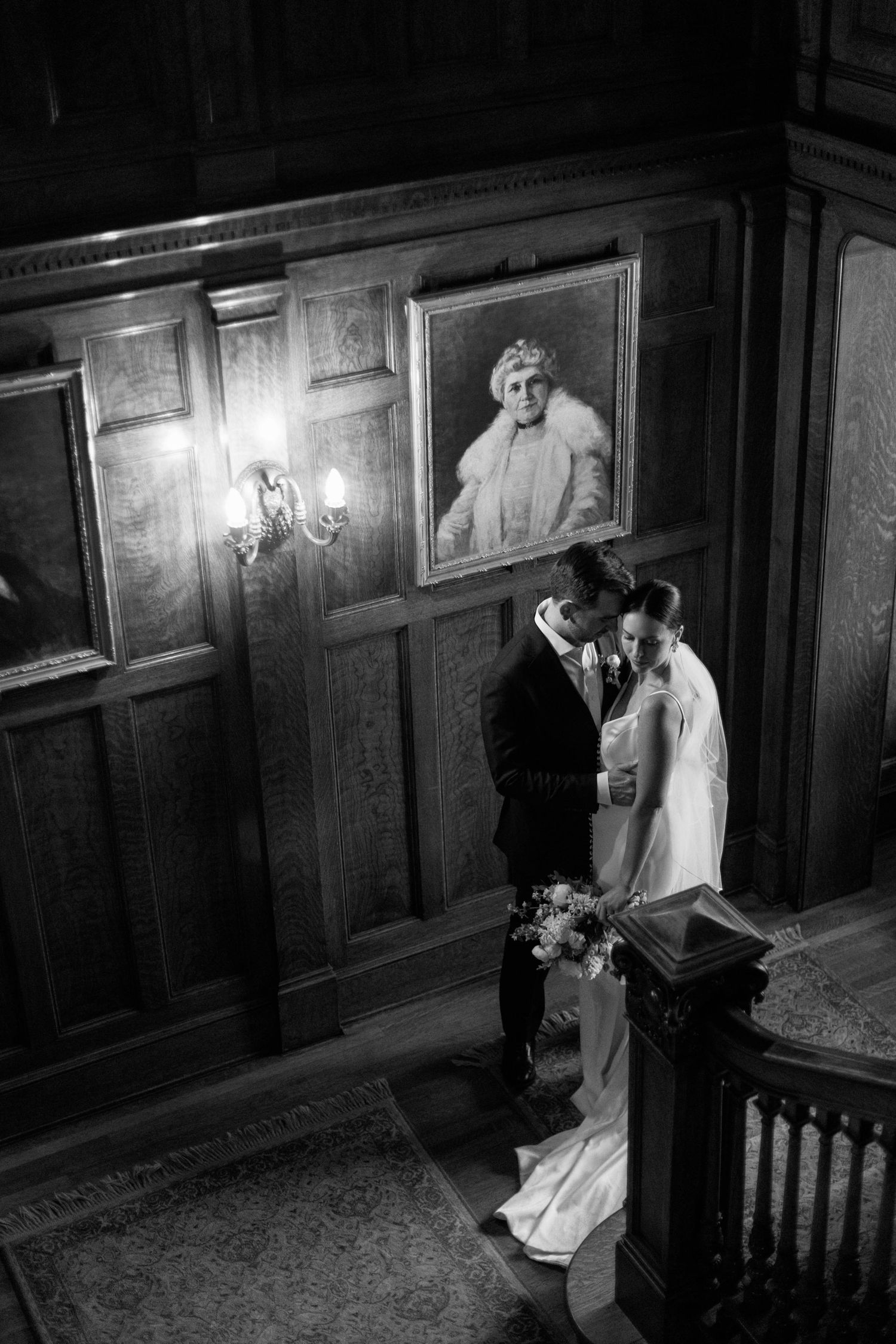 Black and white image of couple sharing intimate moment beneath historic portraits in wood-paneled hall at the historic mccune mansion.