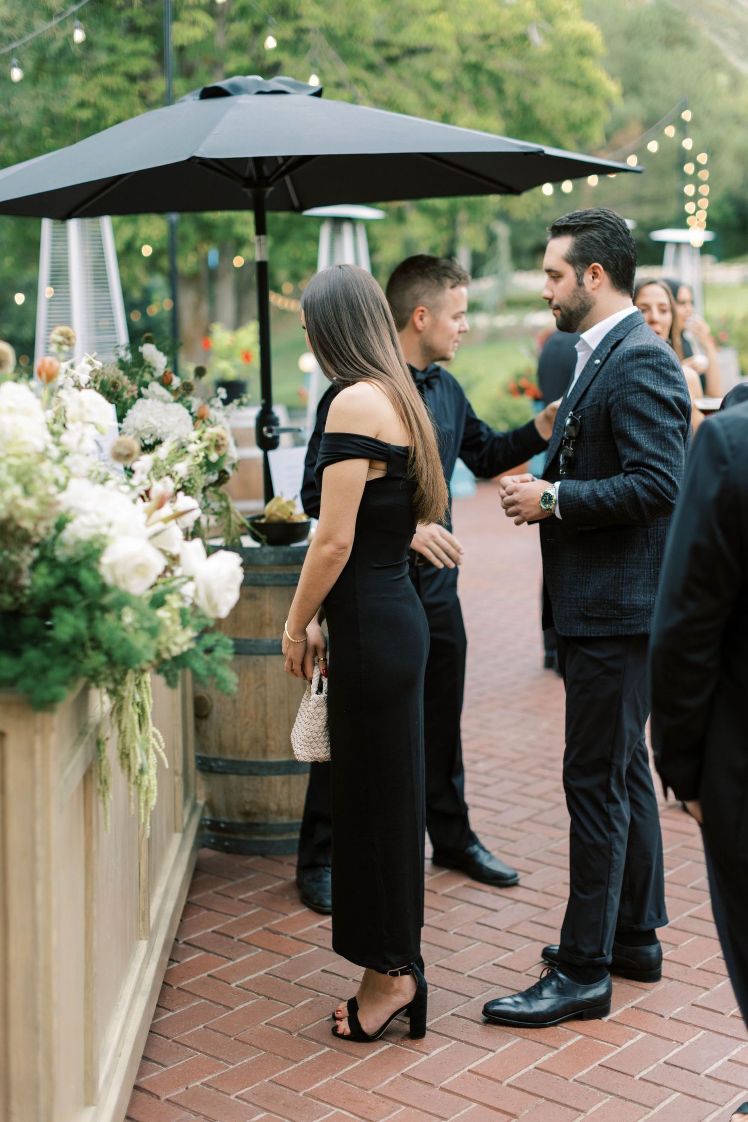 Elegant outdoor cocktail party with guests mingling under black umbrellas and white floral arrangements.
