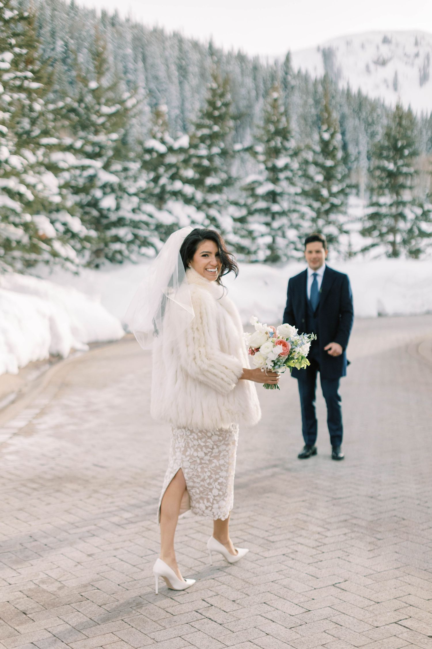Winter wedding photograph on snow-covered mountain path with evergreen trees.