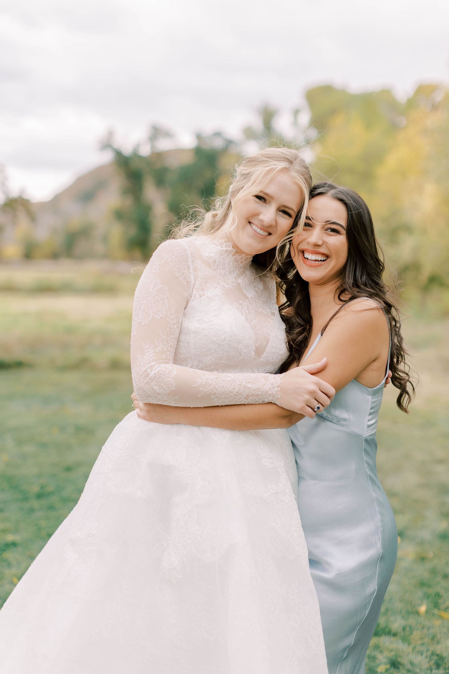 A bride in white lace gown shares a happy moment with bridesmaid in light blue dress.