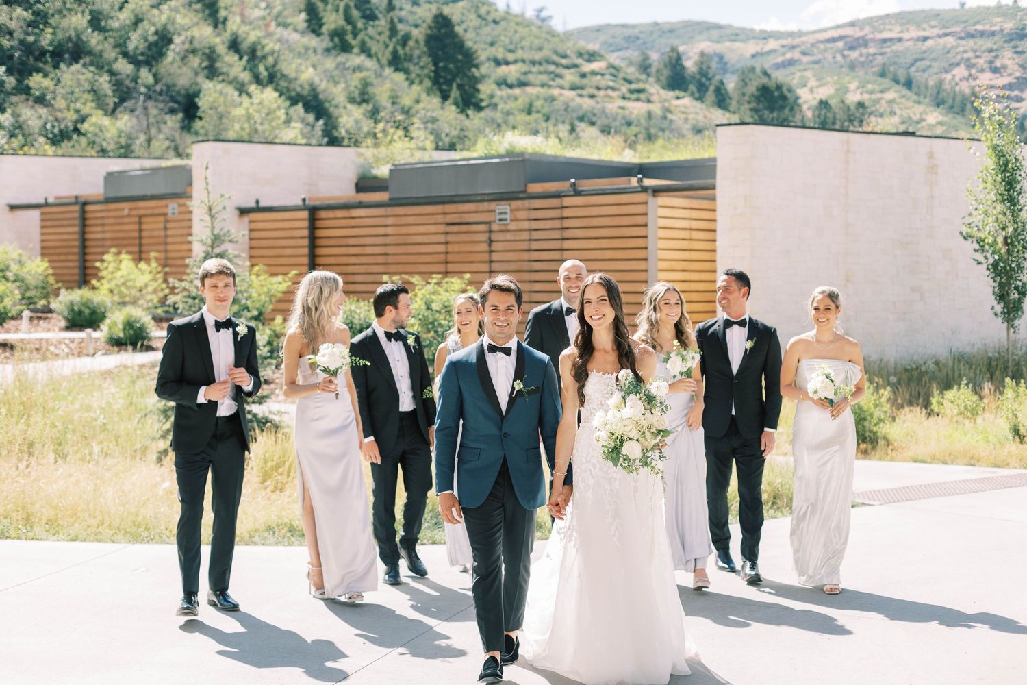 Wedding party walking outdoors with mountain backdrop, featuring bridesmaids and groomsmen in formal attire.