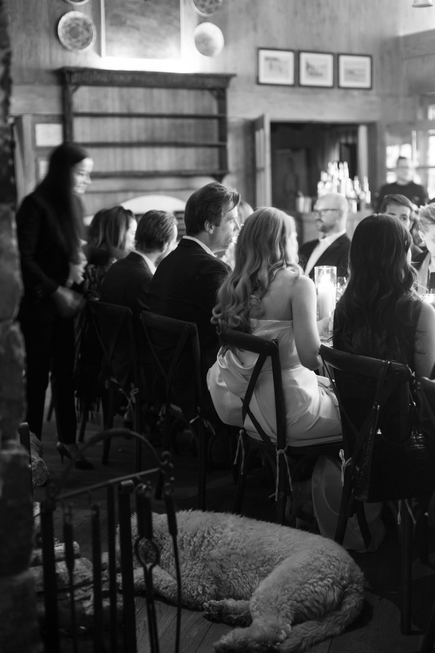 Black and white image of wedding guests seated at tables with a dog resting beneath.